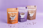 Three BakerCrate mug cake pouches in orange, purple, and brown stand on a lavender background with scattered chocolate sprinkles. 