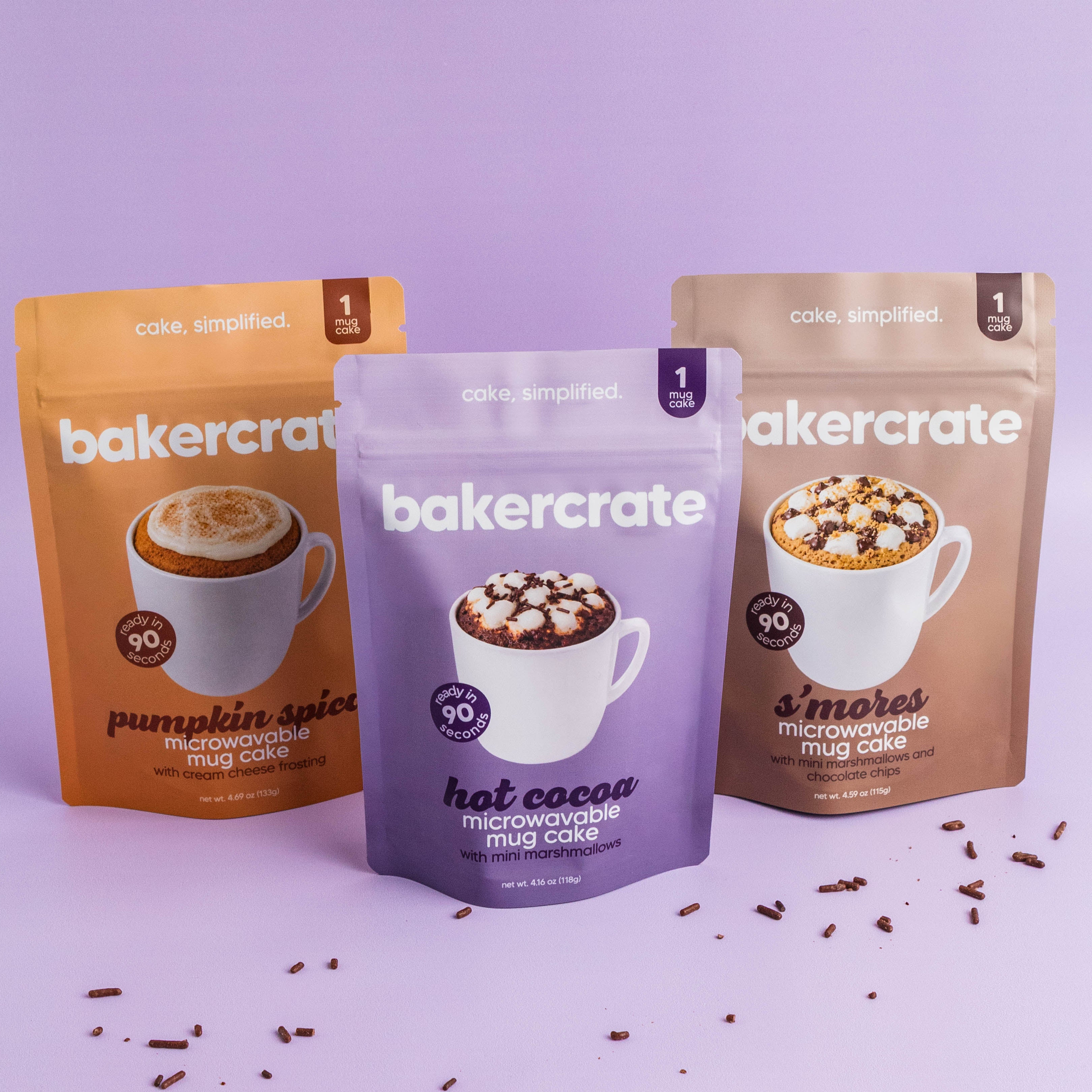 Three bakercrate mug cake pouches, pumpkin spice, hot cocoa and s'mores, on a lavender background with chocolate sprinkles