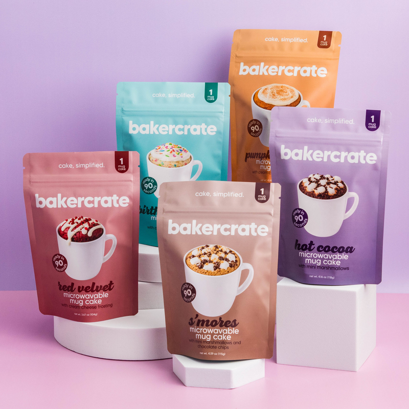 Five Baker Crate mug cake mix pouches in assorted flavors arranged on white display blocks against a pastel purple background. The pouches are in red, teal, orange, brown, and purple packaging.