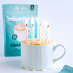 White mug with birthday mug cake and birthday candles next to a BakerCrate birthday cake mug cake mix pouch on a white background