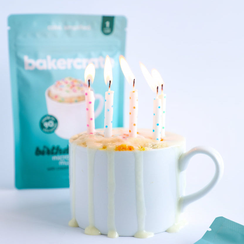 White mug with birthday mug cake and candles next to a BakerCrate mug cake mix ouch on a white background