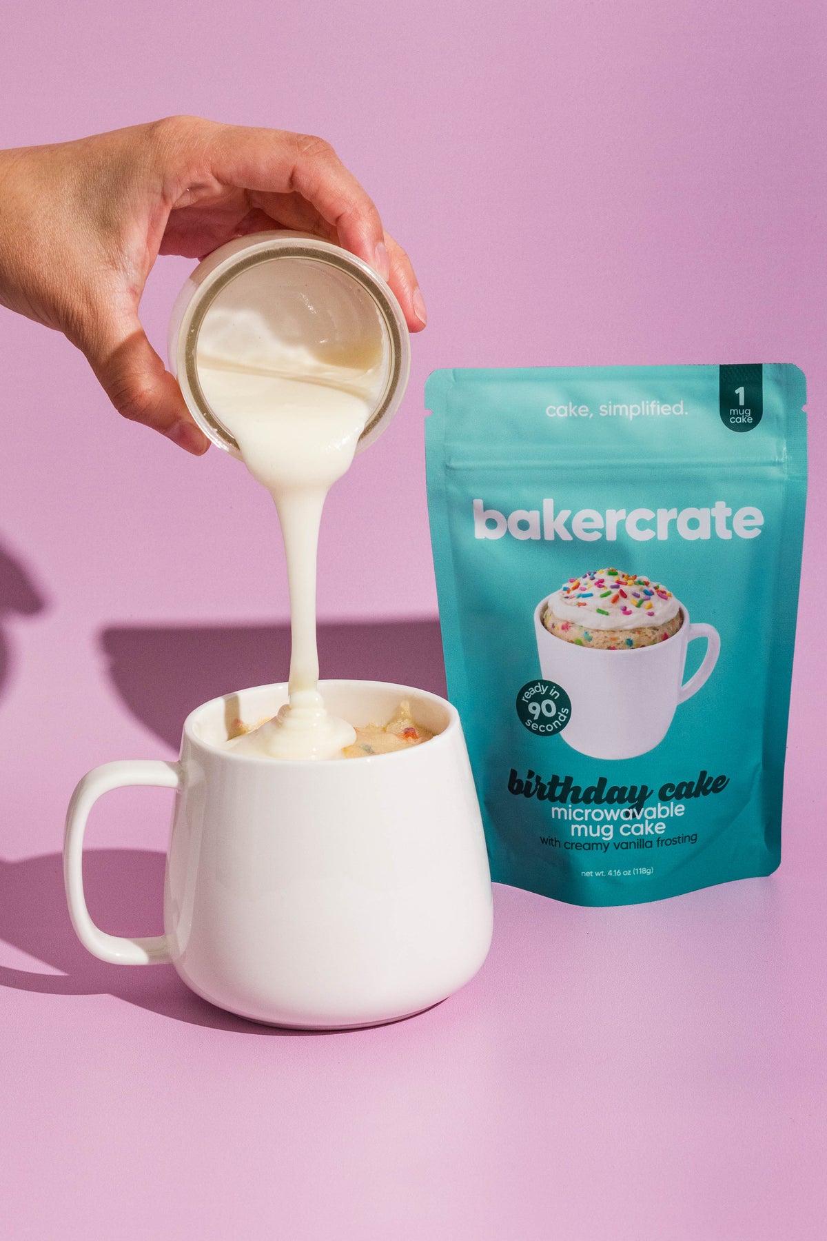 A hand pours a thick white liquid into a white ceramic mug next to a teal stand-up pouch of bakercrate Birthday Cake Microwavable Mug Cake on a solid pink background.