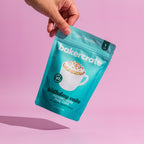 A hand holds a teal stand-up pouch of Bakercrate birthday cake microwavable mug cake mix against a solid pink background.