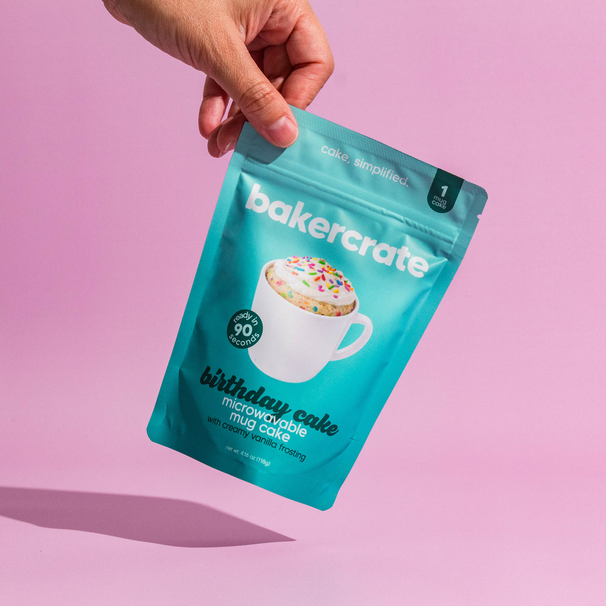 A hand holds a teal stand-up pouch of Bakercrate birthday cake microwavable mug cake mix against a solid pink background.