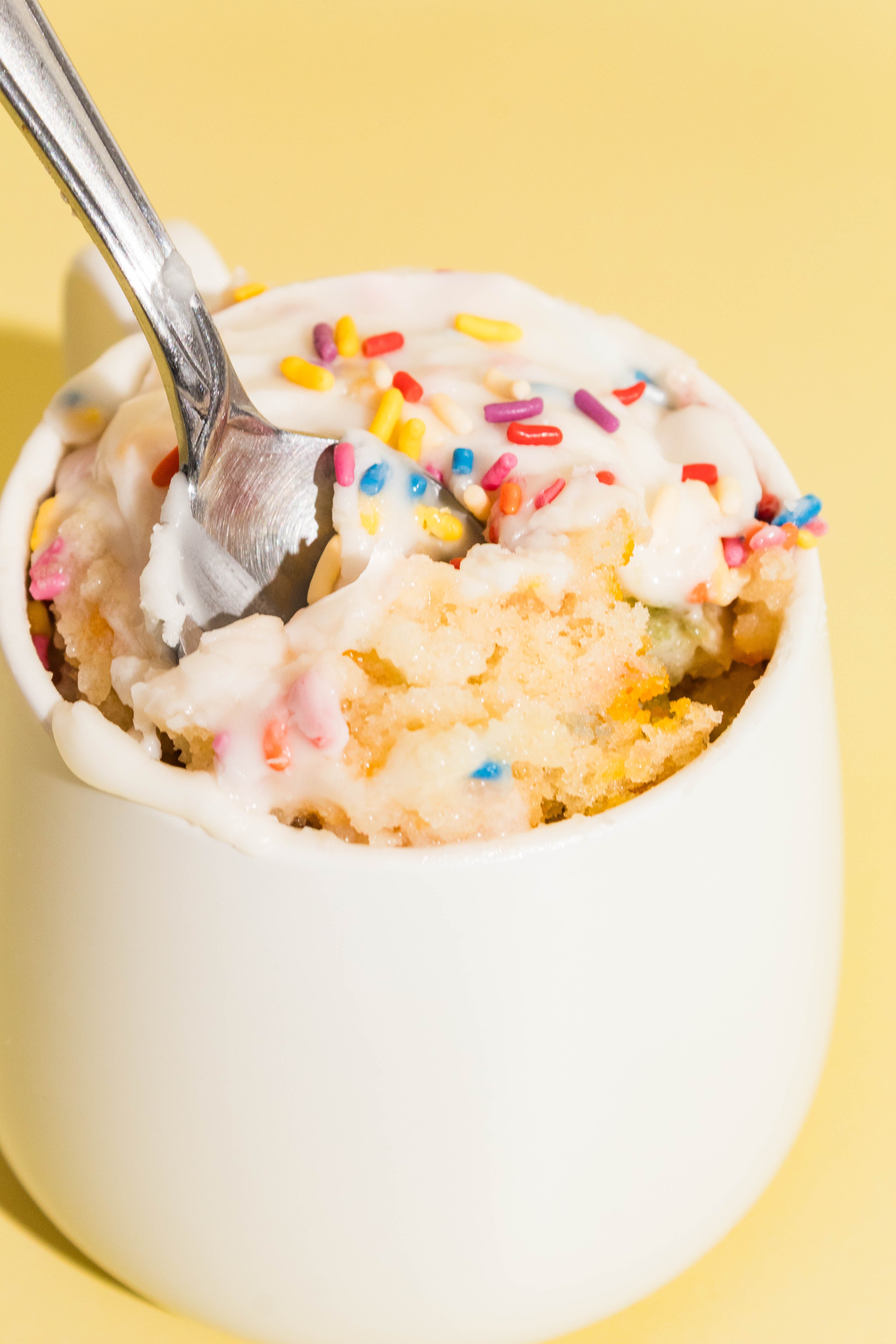 Close-up of a spoon lifting a bite of birthday cake mug cake on a yellow background