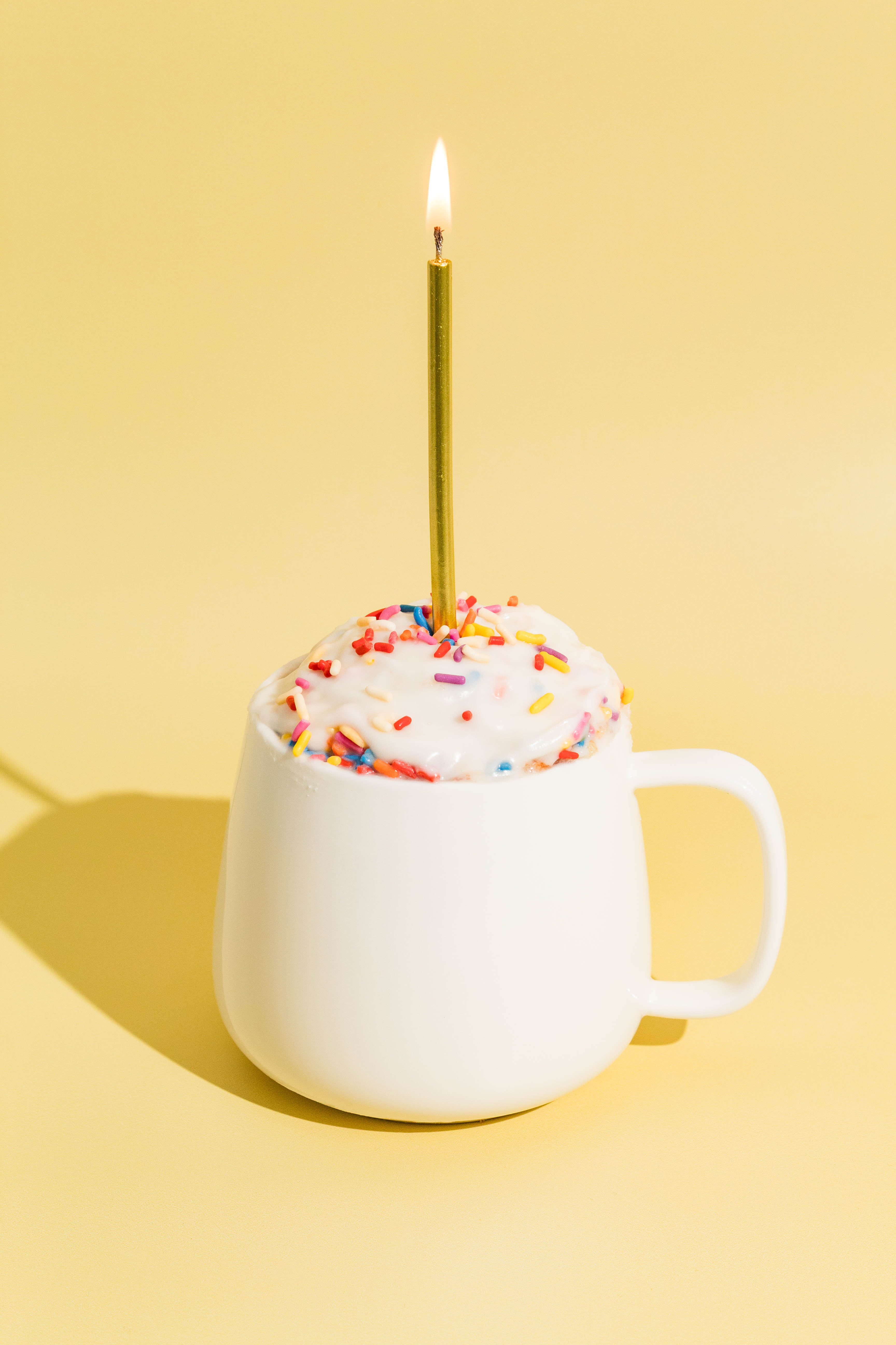 Birthday cake mug cake in a white mug with a lit birthday candle on a yellow background