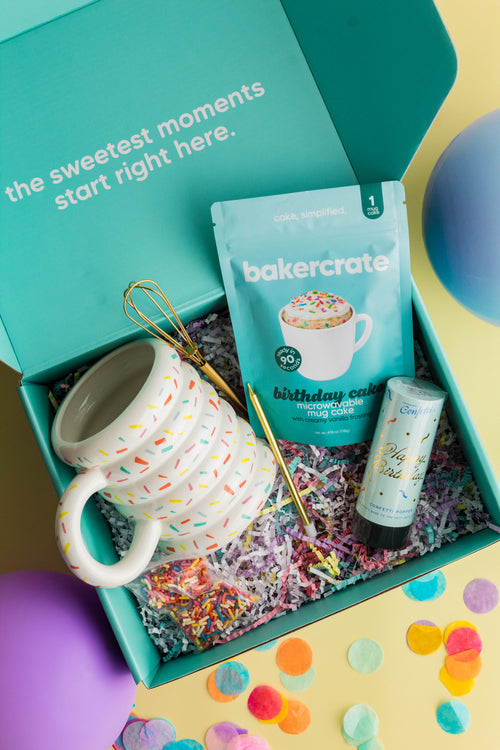 Open BakerCrate Birthday Mug Cake Kit teal gift box with contents: sprinkle-print ceramic mug, gold mini whisk, birthday cake mug cake mix, frosting packet, and confetti popper on a yellow background