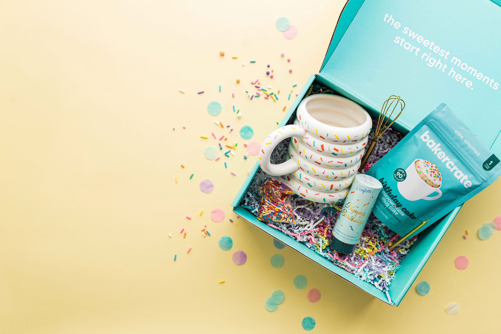A turquoise gift box on a pale yellow background, open to reveal a white ceramic mug with colorful sprinkles, a blue bag of bakerate microwavable mug cake mix, a small confetti popper, sprinkles, a gold whisk, and shredded paper filler with confetti scattered around.