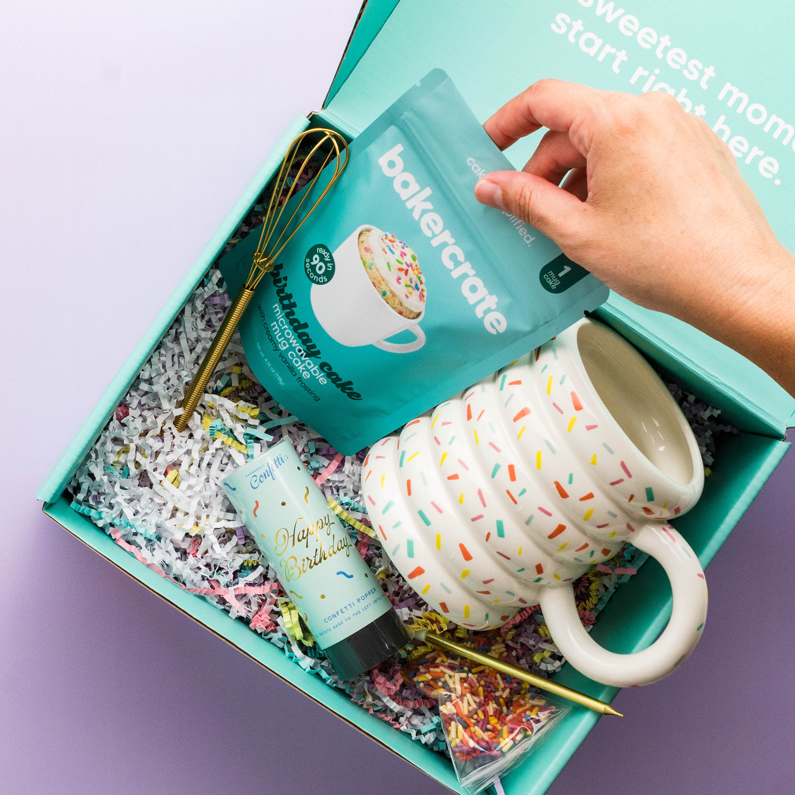 Hand reaching into an open BakerCrate Birthday Mug Cake Kit teal box with birthday cake mug cake mix pouch, ceramic mug, gold mini whisk, and confetti popper on a lavender background
