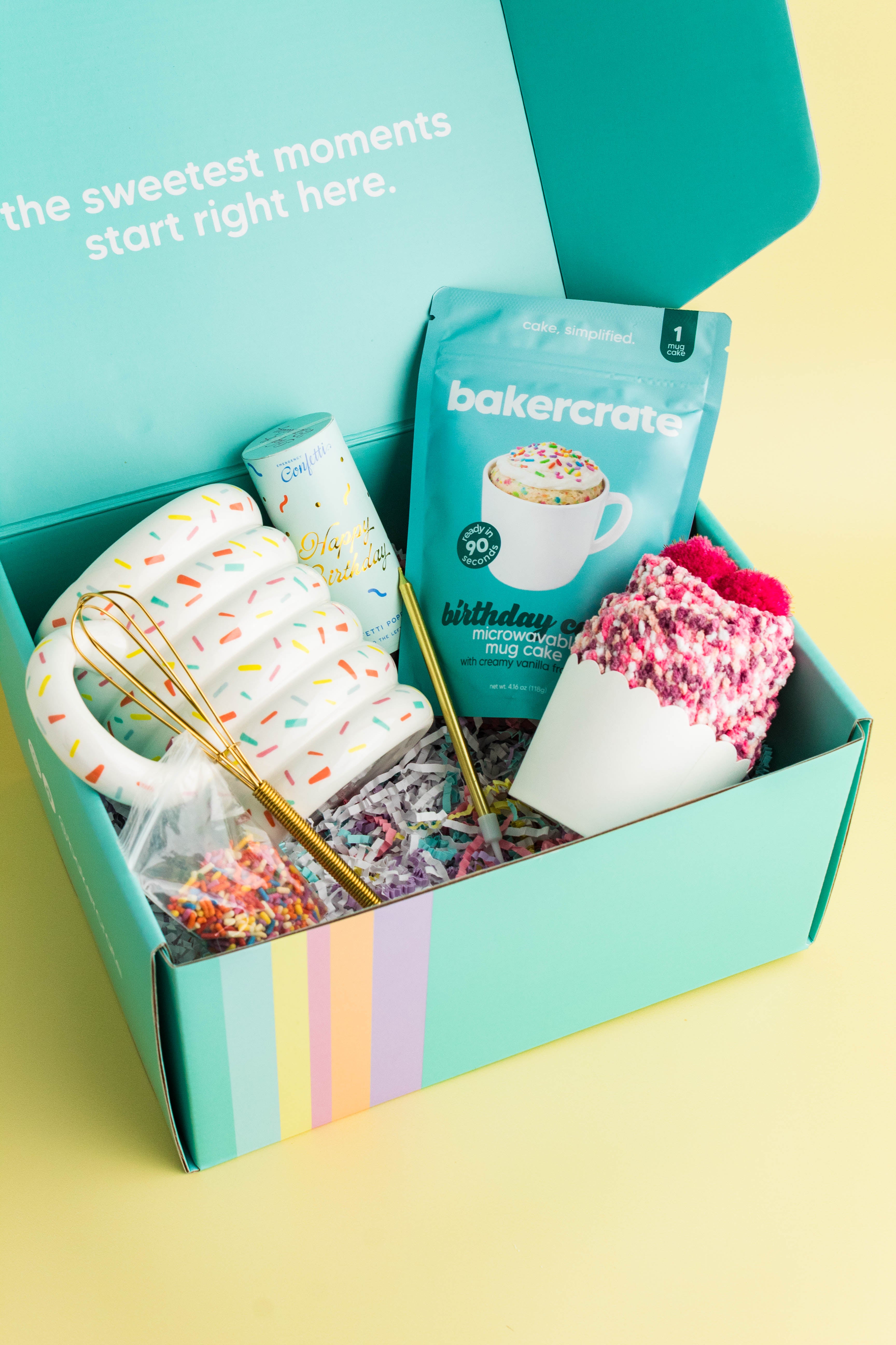 Open BakerCrate Birthday Mug Cake Kit box showing sprinkle-print mug, birthday cake mix, gold mini whisk, and fuzzy birthday socks on a yellow background