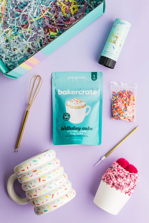 BakerCrate Birthday Mug Cake Kit flatlay showing all contents: mug, birthday cake mix pouch, gold mini whisk, frosting packet, and confetti popper on a lavender background