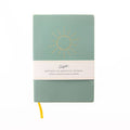 A sage green hardcover journal with a gold sunburst design on the front cover and a white paper band wrapped around the middle. The band reads 'BIRTHDAY CELEBRATION JOURNAL'.