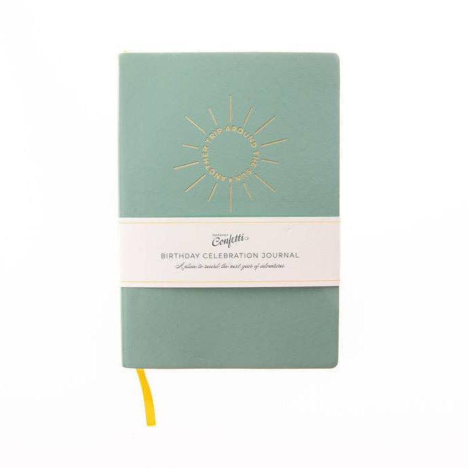 A sage green hardcover journal with a gold sunburst design on the front cover and a white paper band wrapped around the middle. The band reads 'BIRTHDAY CELEBRATION JOURNAL'.