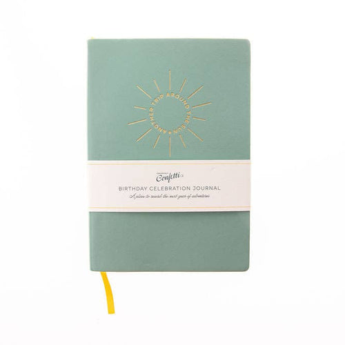 A sage green hardcover journal with a gold sunburst design on the front cover and a white paper band wrapped around the middle. The band reads 'BIRTHDAY CELEBRATION JOURNAL'.