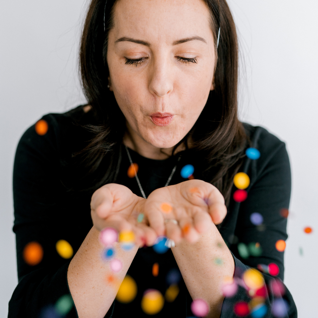 Person blowing a handful of multicolored confetti toward the camera against a white background