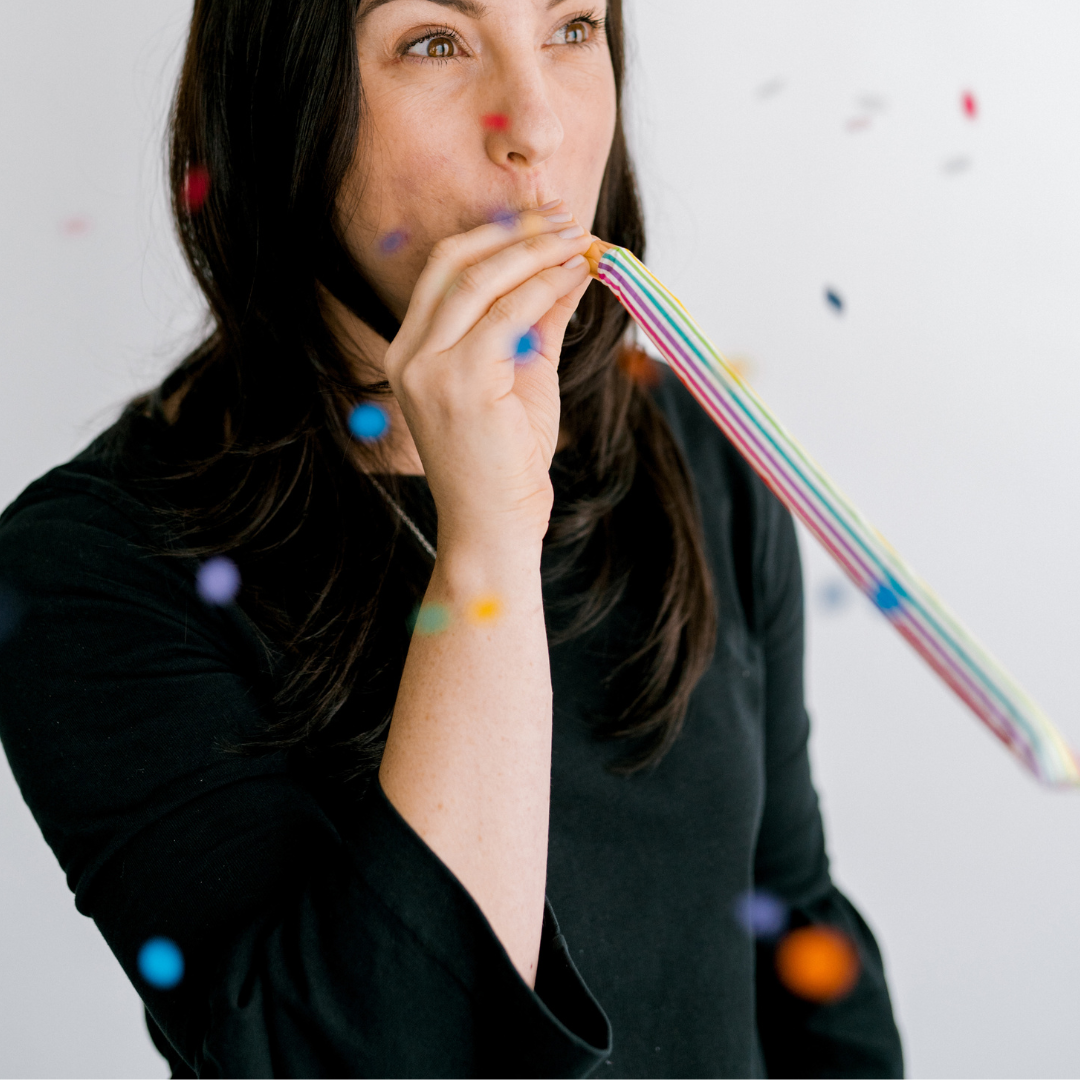 Person blowing a rainbow striped party horn with confetti falling around them