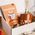 A white gift box contains a bag of Bakercrate pumpkin spice mug cake mix and a terracotta-colored ceramic mug with white leaf patterns and a gold whisk inside. The box is styled with a small orange pumpkin, autumn leaves, and a beige striped blanket on a marble surface.