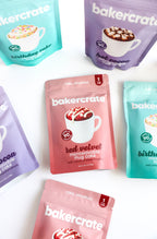 Five BakerCrate mug cake mix pouches in red velvet, hot cocoa, and birthday cake flavors scattered on a white surface