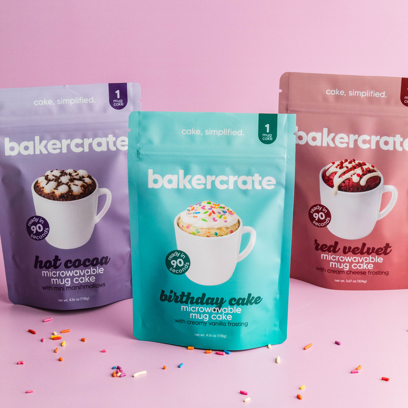 Three Baker Crate microwavable mug cake pouches arranged on a pink background: a purple Hot Cocoa pouch on the left, a teal Birthday Cake pouch in the center, and a dusty pink Red Velvet pouch on the right. Small sprinkles are scattered around the base.