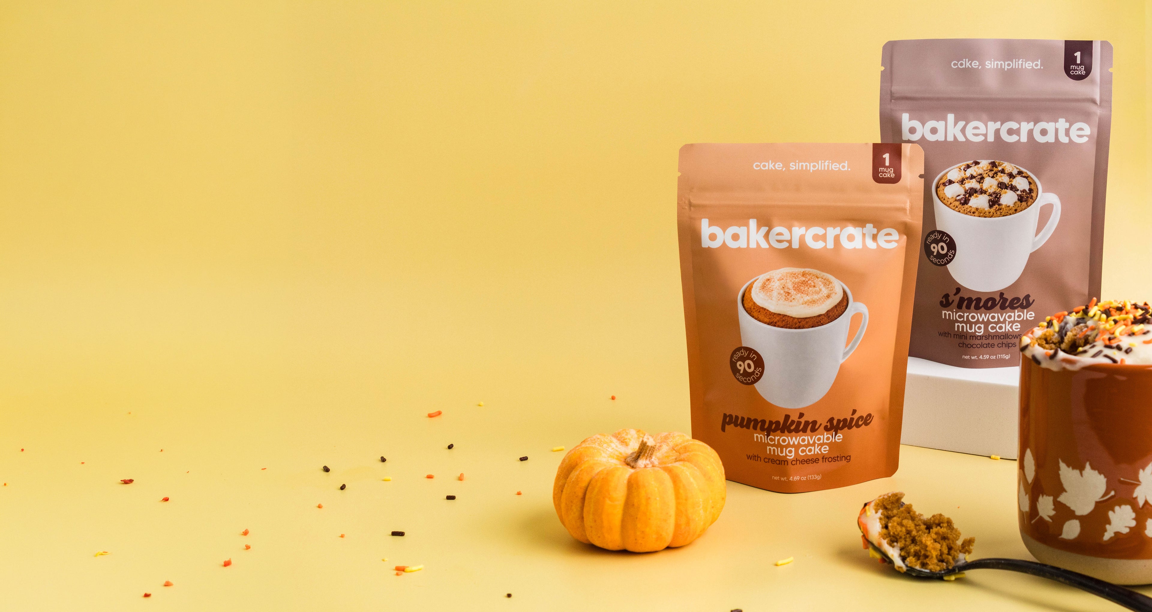 bakercrate pumpkin spice and s'mores mug cake pouches on a yellow background with a mini pumpkin and a spoonful of mug cake