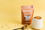 A stand-up pouch of bakercrate Pumpkin Spice Microwave Mug Cake mix stands on a yellow studio background, with a white mug filled with colorful cookie crumbs and a spoonful of cake in front. The package is tan and white with a pumpkin spice illustration.