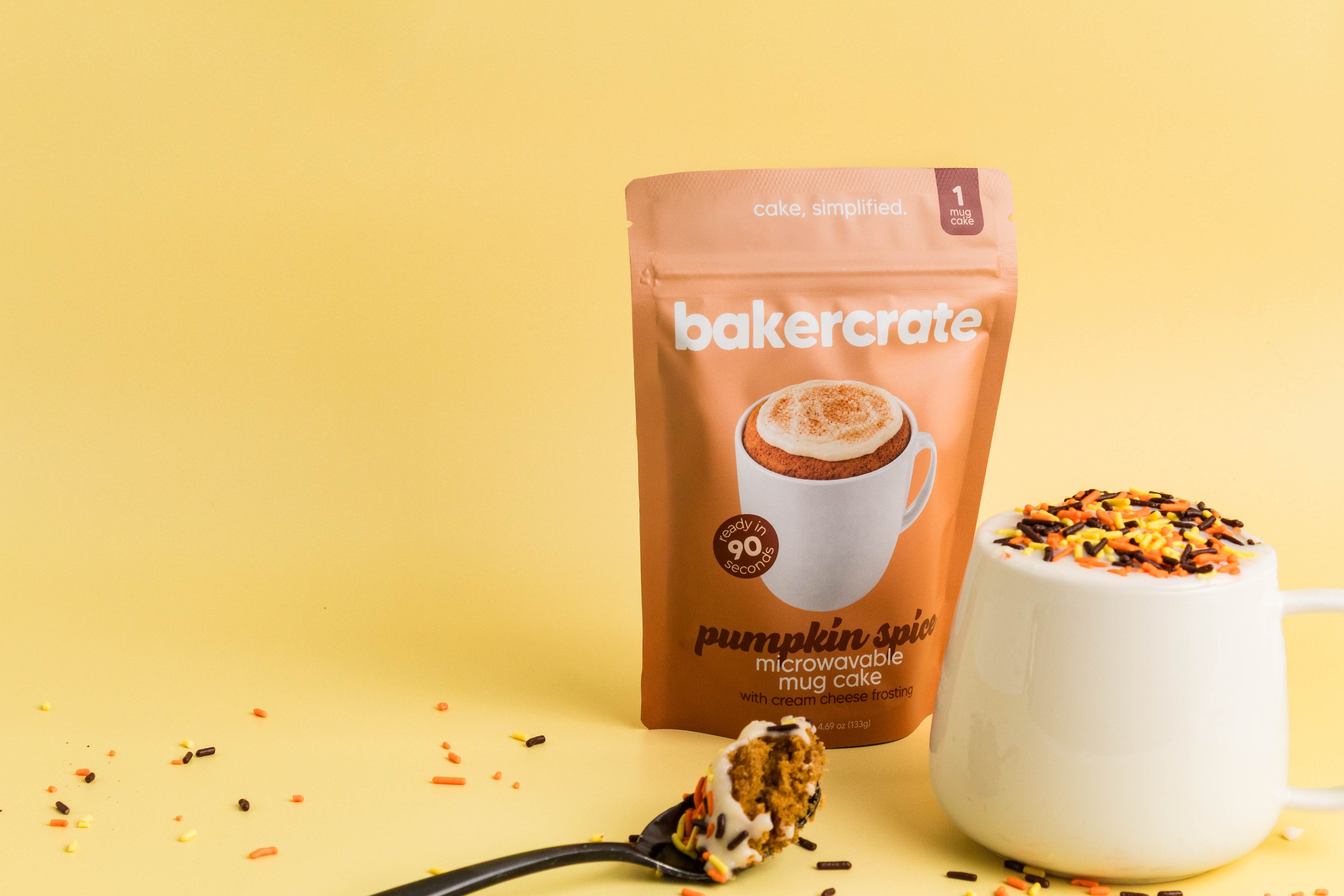A stand-up pouch of bakercrate Pumpkin Spice Microwave Mug Cake mix stands on a yellow studio background, with a white mug filled with colorful cookie crumbs and a spoonful of cake in front. The package is tan and white with a pumpkin spice illustration.