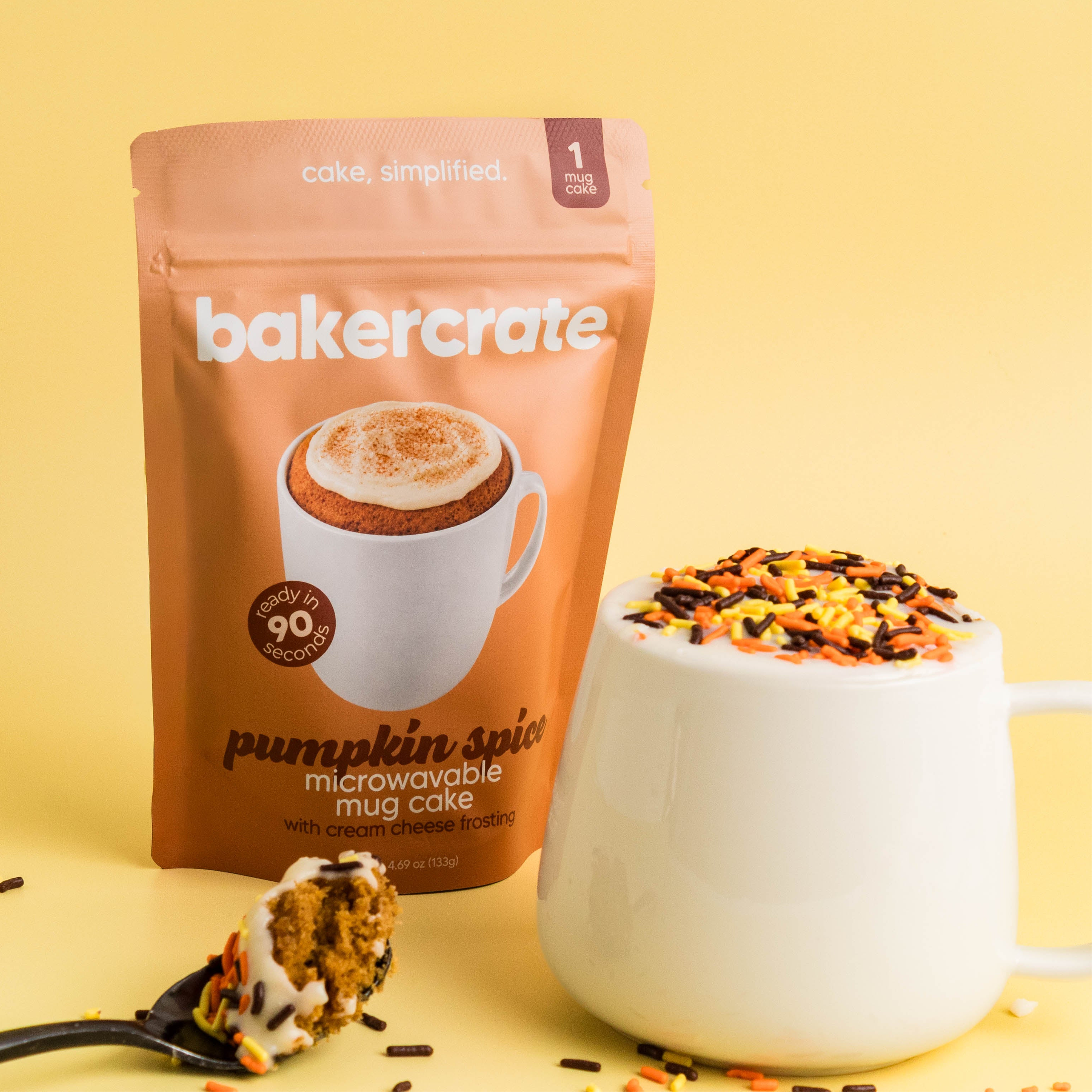 bakercrate pumpkin spice mug cake pouch beside a frosted mug cake with autumn sprinkles and a spoonful