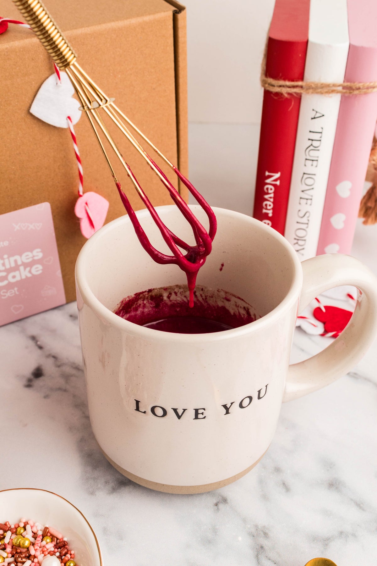 Gold mini wire whisk lifting red velvet batter from a LOVE YOU mug beside a Valentine's gift box
