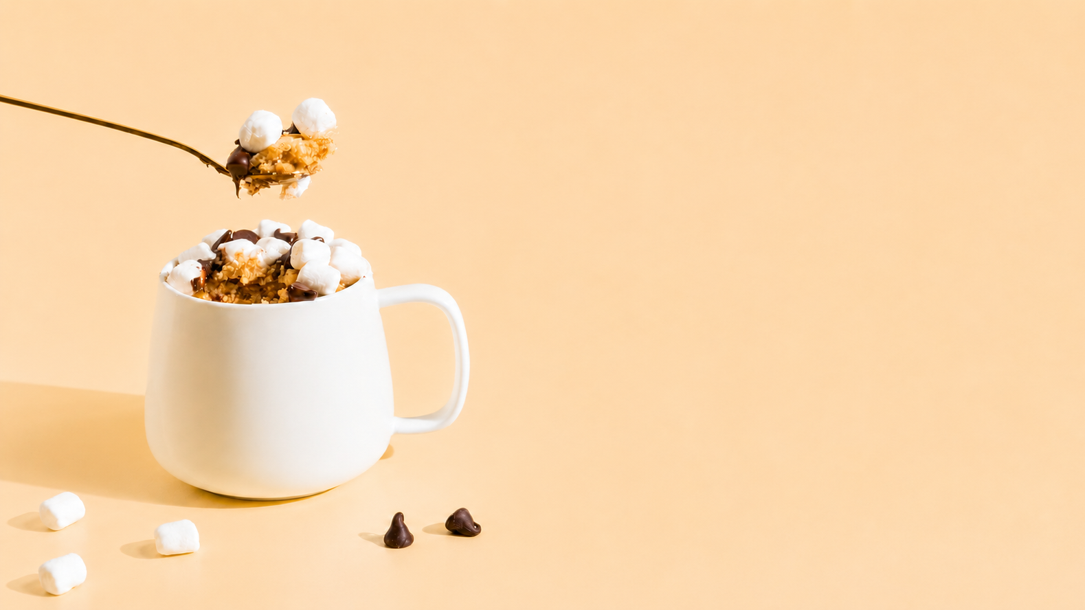 A white ceramic mug filled with a light brown mug cake with marshmallows and chocolate pieces, with a spoon lifting a scoop above it, set against a solid warm peach background.
