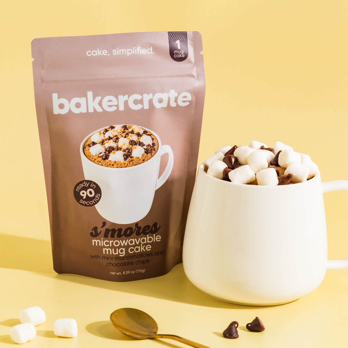 A bag of bakercrate S'mores Microwavable Mug Cake stands next to a white ceramic mug filled with marshmallows and chocolate chips on a warm yellow background.