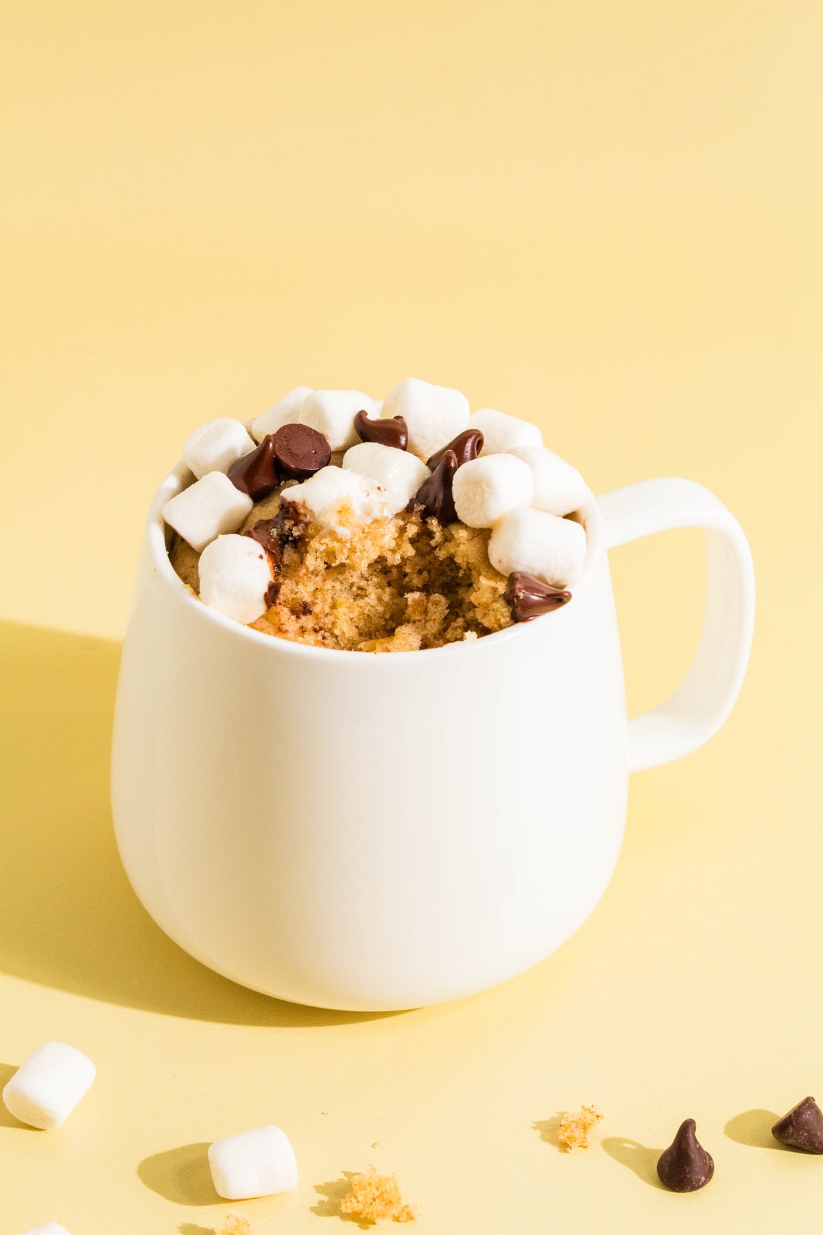 S'mores mug cake topped with mini marshmallows and chocolate chips in a white mug on a yellow background
