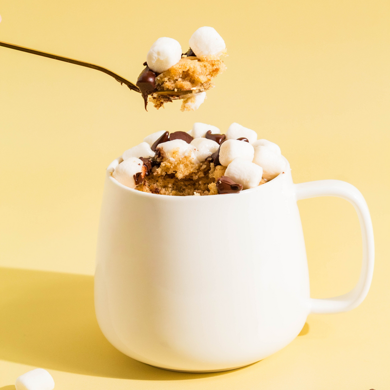 A white ceramic mug filled with a s'mores flavored mug cake topped with mini marshmallows and chocolate chips, with a spoon lifting a portion above the mug, set against a solid pale yellow background.