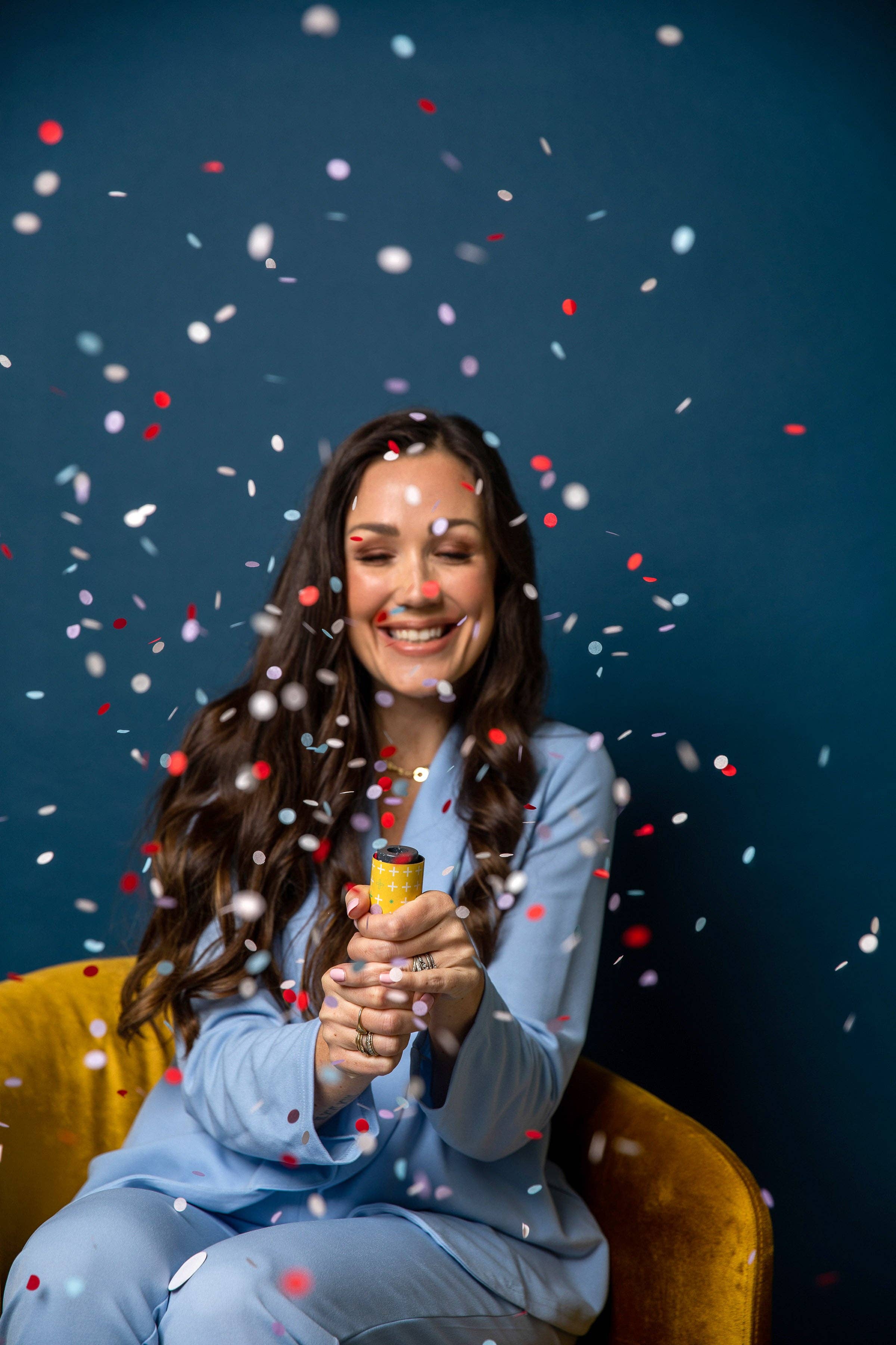 Person seated in a mustard velvet chair setting off a yellow confetti popper, with confetti falling around them