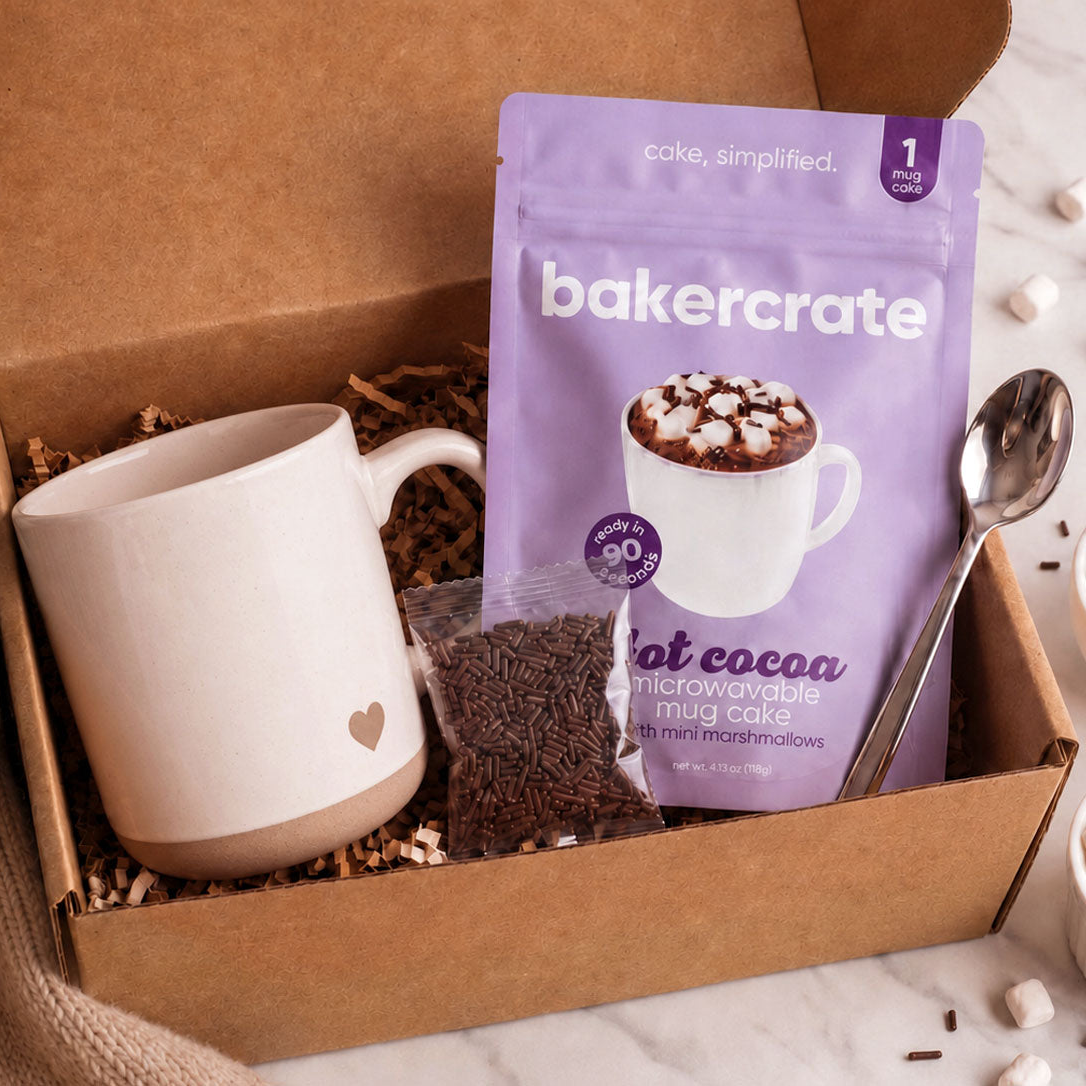 Open kraft gift box with a heart-embossed stoneware mug, Bakercrate hot cocoa microwavable mug cake pouch, chocolate sprinkles packet, and silver spoon on shredded paper