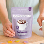 Hands holding a Bakercrate hot cocoa mug cake mix pouch on a wooden cutting board