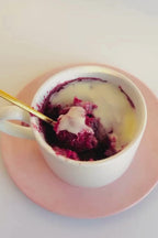 Gold spoon lifting a bite of red velvet mug cake with cream cheese frosting from a white mug on a pink saucer