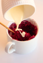Cream cheese frosting being poured over red velvet mug cake in a white mug