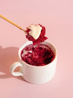 Red velvet mug cake with cream cheese frosting in white mug, gold spoon, pink background