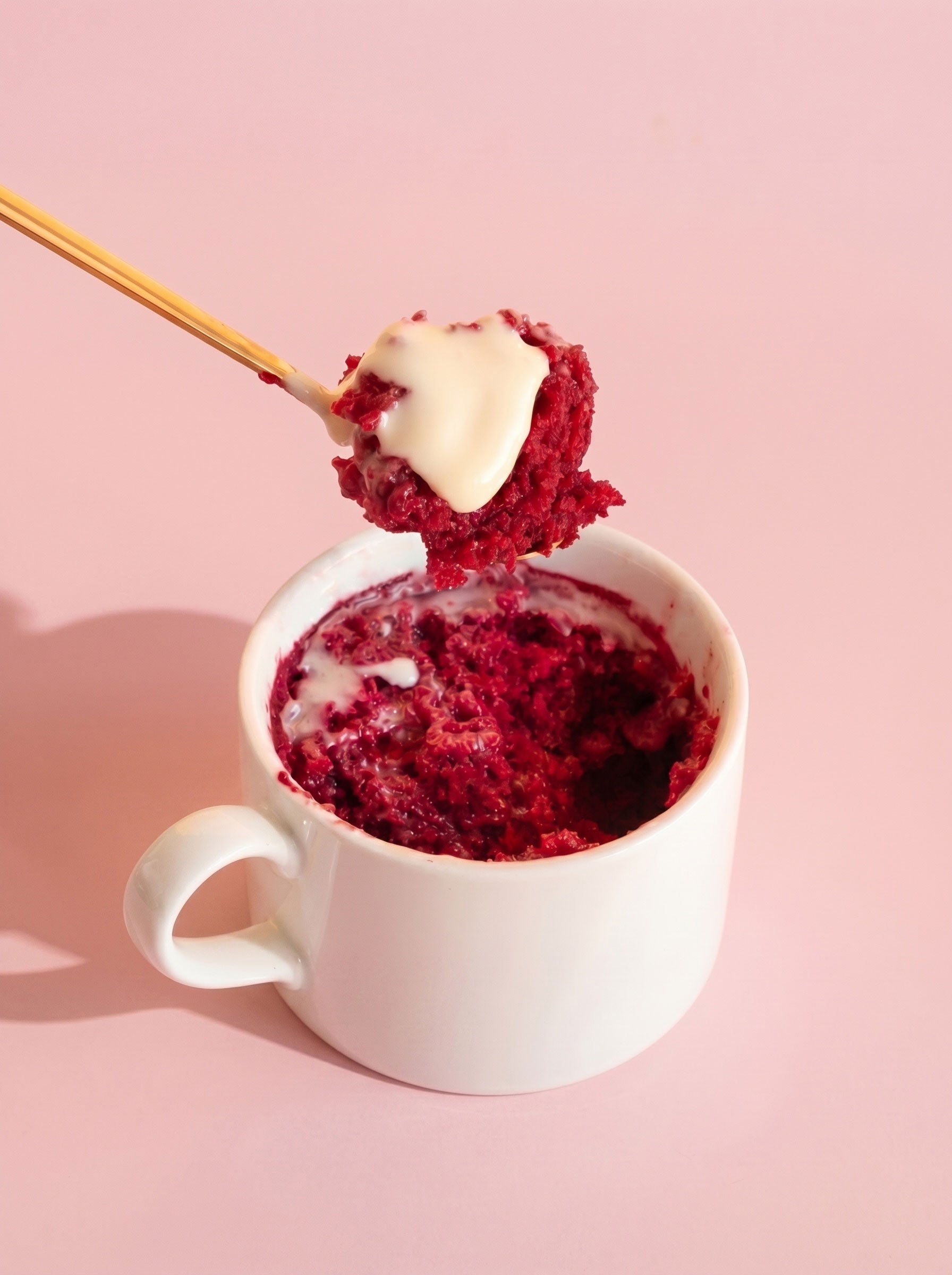Red velvet mug cake with cream cheese frosting in white mug, gold spoon, pink background