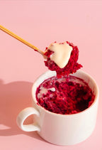 Red velvet mug cake with cream cheese frosting and gold spoon on pink background