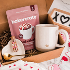 Open kraft gift box with a heart-embossed stoneware mug, Bakercrate red velvet microwavable mug cake pouch, sprinkles vial, and gold spoon on a heart-shaped dish with Valentine's confetti
